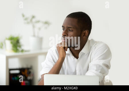 Young african-american thoughtful businessman holding hand on chin planning future project at work with laptop, black meditative african entrepreneur Stock Photo