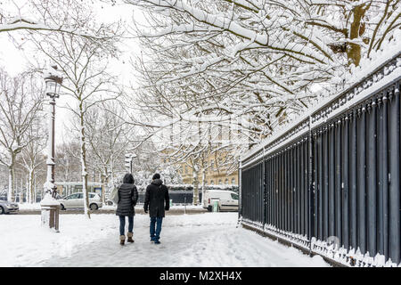 Winter in Paris in the snow. Two people wrapped up in warm clothes walk on a snow covered sidewalk along a tall black fence in a chic neighborhood. Stock Photo
