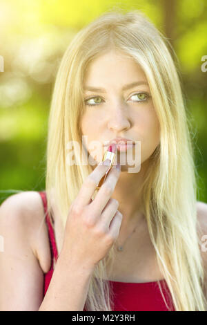 Beautiful young blonde woman corrects-applying red lipstick make-up in the park.  Concept teenager lifestyle. Stock Photo