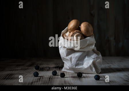 Horizontal photo with several brown edible mushrooms. Raw champignons are in white paper bag. Bag is on vintage wooden board with spilled blackthorns. Stock Photo