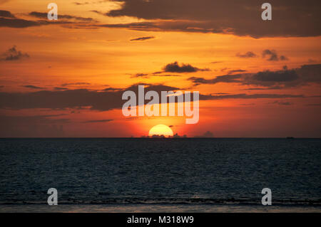 Huge orange semicircle of the sun sets behind the water, the red sky with gray clouds, the sunset in the Maldives. Stock Photo
