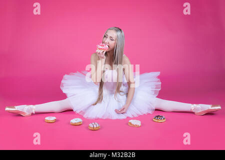 Pretty blonde ballerina eats a donut sitting in a split in tutu skirt. Stock Photo