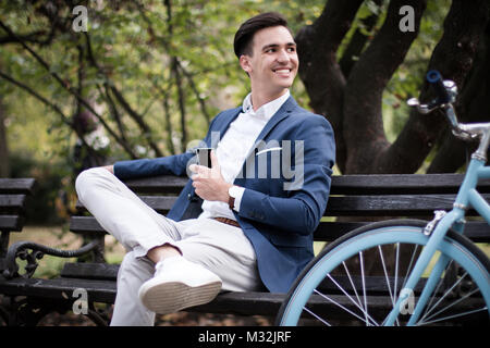 Young businessman with smartphone sitting on a bench in a park, with his bicycle beside him. Stock Photo
