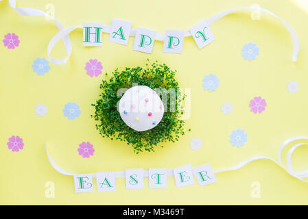 Top view holiday composition with green sprouts pillow and decorated easter cupcake, Happy Easter lettering with white ribbon on bright yellow backgro Stock Photo