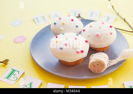 Plate with delicious Easter cupcakes and vintage ribbon with Happy Easter lettering, fresh branches with kidneys and paper flowers on bright yellow ba Stock Photo