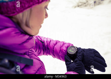 Woman hiker checking the elevation on sports watch, smartwatch with altimeter app in winter woods and mountains. Female trekker in white snowy forest  Stock Photo