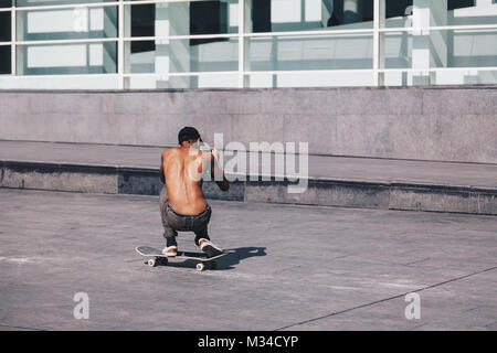 Skater taking photo or gaming with his smartphone during skating with blurred modern structure on the background. Stock Photo