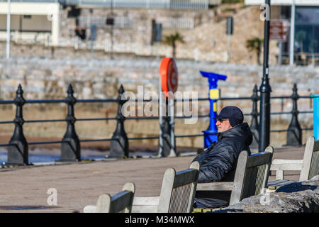 Older man sat on bench on Torquay promenade, in bright sunny but chilly weather. Torquay, Torbay, Devon, UK. February 2018. Stock Photo