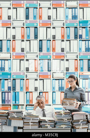 Stressed businessman working at office desk and overloaded with work, the desktop is covered with paperwork, his secretary is bringing more files Stock Photo