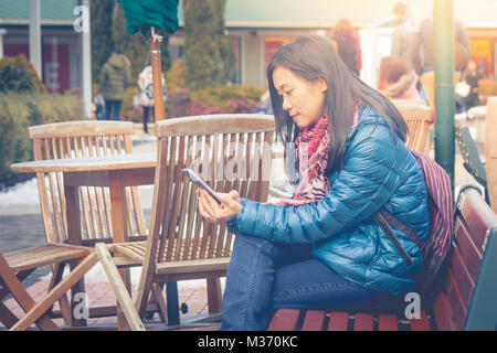 Travel winter vacation concept : Asian woman traveler sitting on wooden long bench at outside and playing her smartphone. Stock Photo