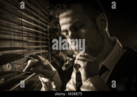 Gangster smoking a cigar while looking out the window at a car headlights Stock Photo