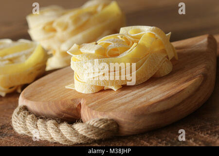Nest of pasta tagliatelle on wooden cutting board aon rustic background. Selective focus Stock Photo