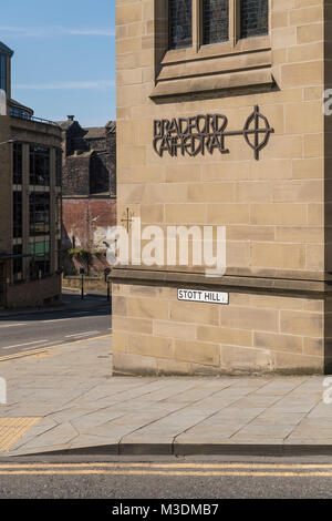 Small corner of Bradford Cathedral with modern name sign on stone wall, at roadside & with old & new buildings beyond - West Yorkshire, England, UK. Stock Photo