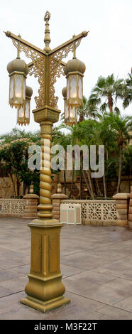Ornate vintage golden illuminated lamp post. Ornamental gate & palm trees in background. At Disneysea Tokyo. Stock Photo