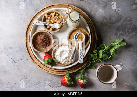 Ingredients for cooking chocolate mug cakes. Flour, cocoa powder, sugar, caramel in wooden bowls, milk, strawberries and mint served with spoons and m Stock Photo