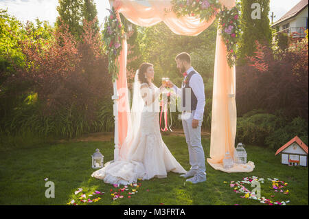 Beautiful couple going to drink from the glasses under the arch outdoors Stock Photo