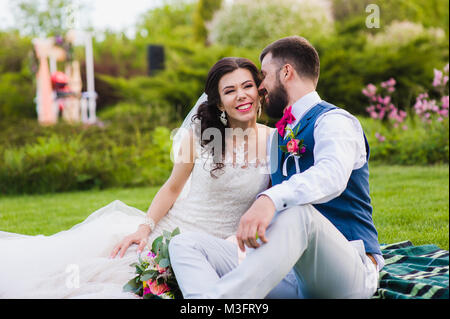 Groom telling something funny to the bride while they and sitting in the green park. Beautiful couple laughing and joking together at their wedding da Stock Photo