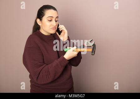 Helpless woman holding hammer with wrench talking on phone hands in a DIY concept Stock Photo