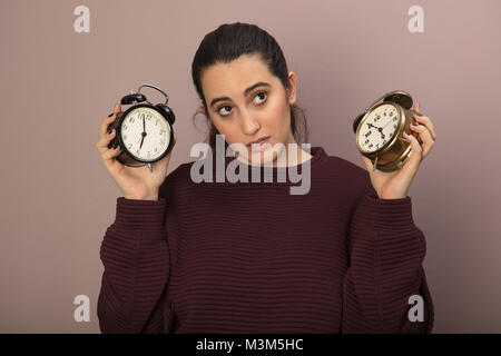 Thoughtful woman holding up two old fashioned alarm clocks with bells showing different times as she looks upwards with a pensive expression trying to Stock Photo