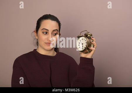 Young woman looking dubiously at an alarm clock with a look of contemplation as she holds it up in her hand Stock Photo