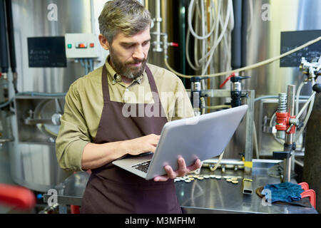 Serious handsome brewer using laptop in factory shop Stock Photo