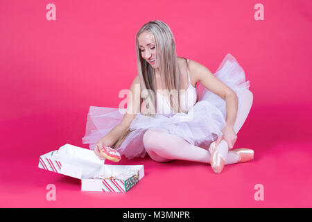 Pretty blonde ballerina in tutu skirt reaching for a donut on pink backdrop. Stock Photo