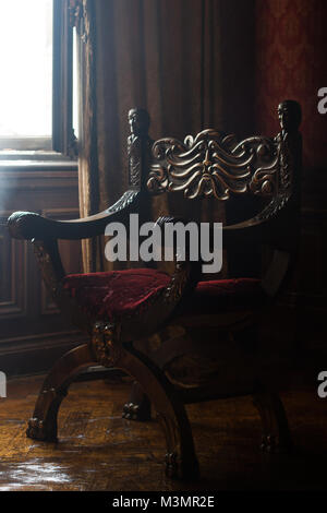 side view of antique armchair with round shape and wooden carved arms in the corner of a classical style room with vintage red wall paper natural ligh Stock Photo