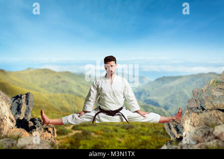 Martial arts karate master in white kimono and black belt doing extreme stretching exercise between the tops of the mountains Stock Photo