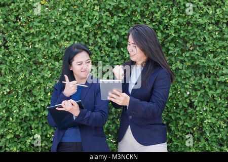 Two asian businesswoman thumbs up for business success outside office at green leaves hedge,Female leadership concept.digtital lifestyle Stock Photo