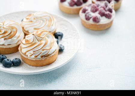 Delicious lemon and raspberry tartlets with meringue on a white vintage plate. Sweet treat on a light blue background. Stock Photo