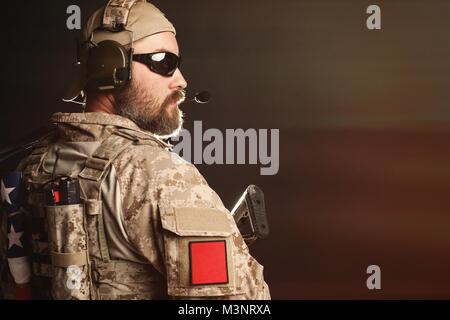 Brutal man in the military desert uniform and body armor is pathetic and looks away at the black background in the Studio. The bearded player in the a Stock Photo