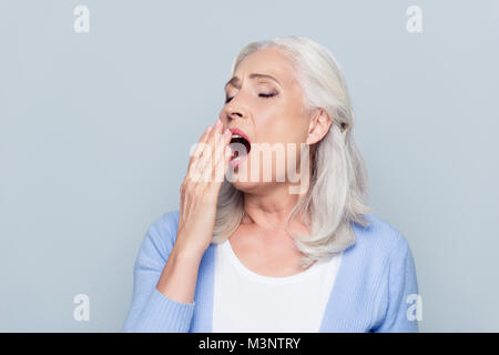 Portrait of tired, bored, attractive, aged, lazy woman in  closing her open mouth with palm, she wants to sleep, yawning over grey background Stock Photo
