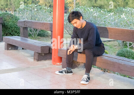 A boy in black clothes sitting in Thai local train station and watching his watch. Stock Photo