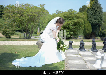 A beautiful dark haired bride wearing a fitted expensive Wedding Dress on a sunny day- brightly lit playing a large chess game Stock Photo
