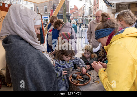 York, UK, 12th February 2018. Woman dressed as a Viking at the annual Jorvik Viking Festival. She is taking part in a realistic re-enactment of life at a Viking market & is standing by an open-air stall. Members of the public (adults & children) are fascinated by  the display of goods she is selling - jewellery made mainly from black jet & amber.They are handling them & examining items closely. City centre of York, North Yorkshire, England, UK. Credit: Ian Lamond/Alamy Live News Stock Photo