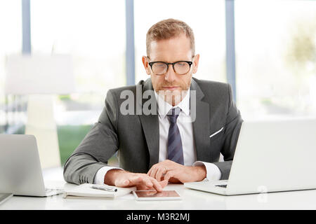 Middle aged financial advisor businessman working on digital tablet while sitting at desk at the office. Stock Photo