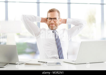 Portrait shot of a successful smiling businessman with hands behind his head sitting at desk with laptops and working at the office. Stock Photo