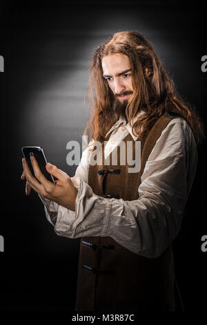 portrait of a man with a beard and long hair wearing a medieval pirate costume on a black background, a pirate holding a mobile phone Stock Photo