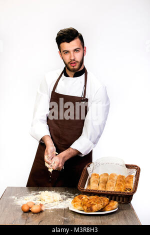 a man in an apron prepares the dough for baking, Cook model on a white background kneads dough on the table decorated with bakery products Stock Photo
