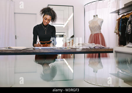 Female fashion designer sketching a design sitting at her table. Fashion entrepreneur engrossed in making a drawing at her desk in her cloth shop. Stock Photo