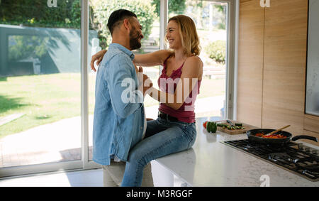 Woman sitting on kitchen counter with man standing. Couple looking at each other and smiling in their kitchen at home. Stock Photo