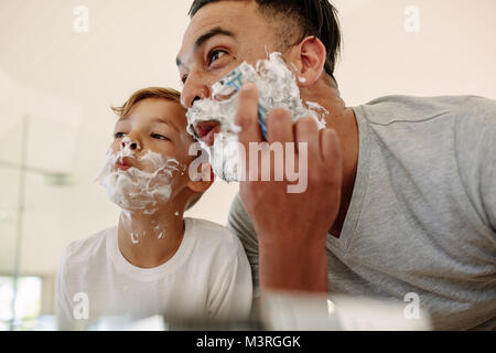Father and son  making funny faces while shaving in bathroom. Young man and little boy with shaving foam on their faces are shaving and looking into t