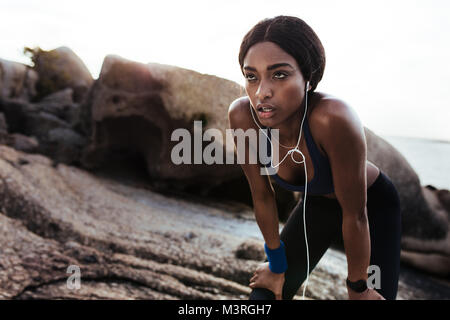 Tired young woman resting after running outdoors. African female runner standing with hands on knees. Stock Photo