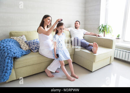 Adorable young pregnant family in living room. Mother combing her daughter's hair. Happiness and love concept Stock Photo