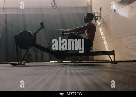 Male using rowing machine at fitness club. Young man doing exercises on fitness machine in gym. Side view. Stock Photo