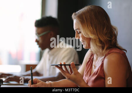 Woman entrepreneur writing in notepad while talking over mobile phone. Man working on laptop sitting at a coffee shop. Stock Photo