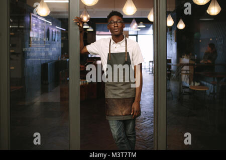 Coffee shop owner standing at the entrance of his shop. Barista standing at a café wearing apron. Stock Photo