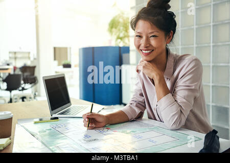 Smiling young Asian architect sitting alone at her desk in a modern office working on a building design Stock Photo