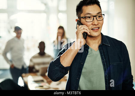 Young Asian designer standing in an office talking on a cellphone with colleagues working in the background Stock Photo