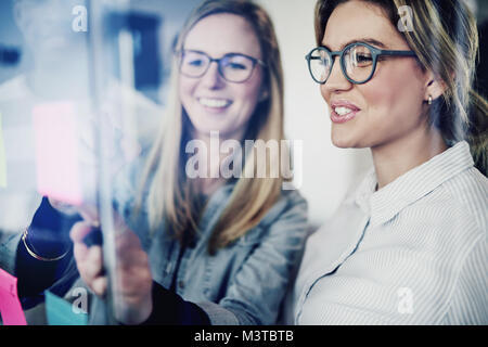 Two young businesswomen standing in a modern office brainstorming together with sticky notes on a glass wall Stock Photo